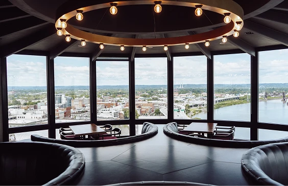 Spacious lounge with curved seating and large windows offering a panoramic view of a cityscape, river, and distant hills. Circular ceiling lights add modern ambiance, emphasizing the scenic outlook.