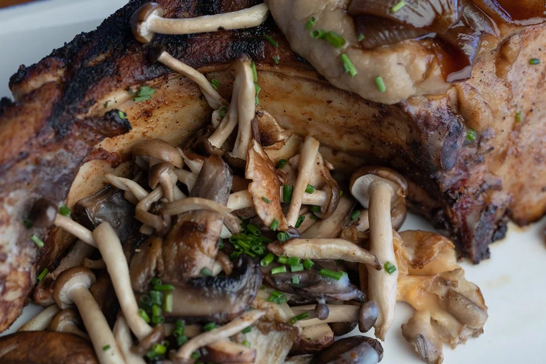 Close-up of a grilled pork chop served with a variety of sautéed wild mushrooms and garnished with chopped chives on a white plate.