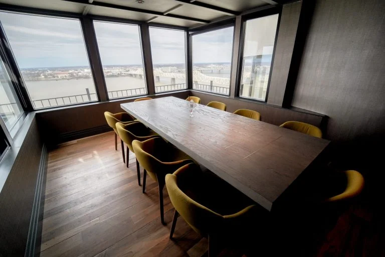 A modern conference room with a wooden table, yellow chairs, and large windows overlooking a river and cityscape. The natural light highlights the minimal, stylish interior.