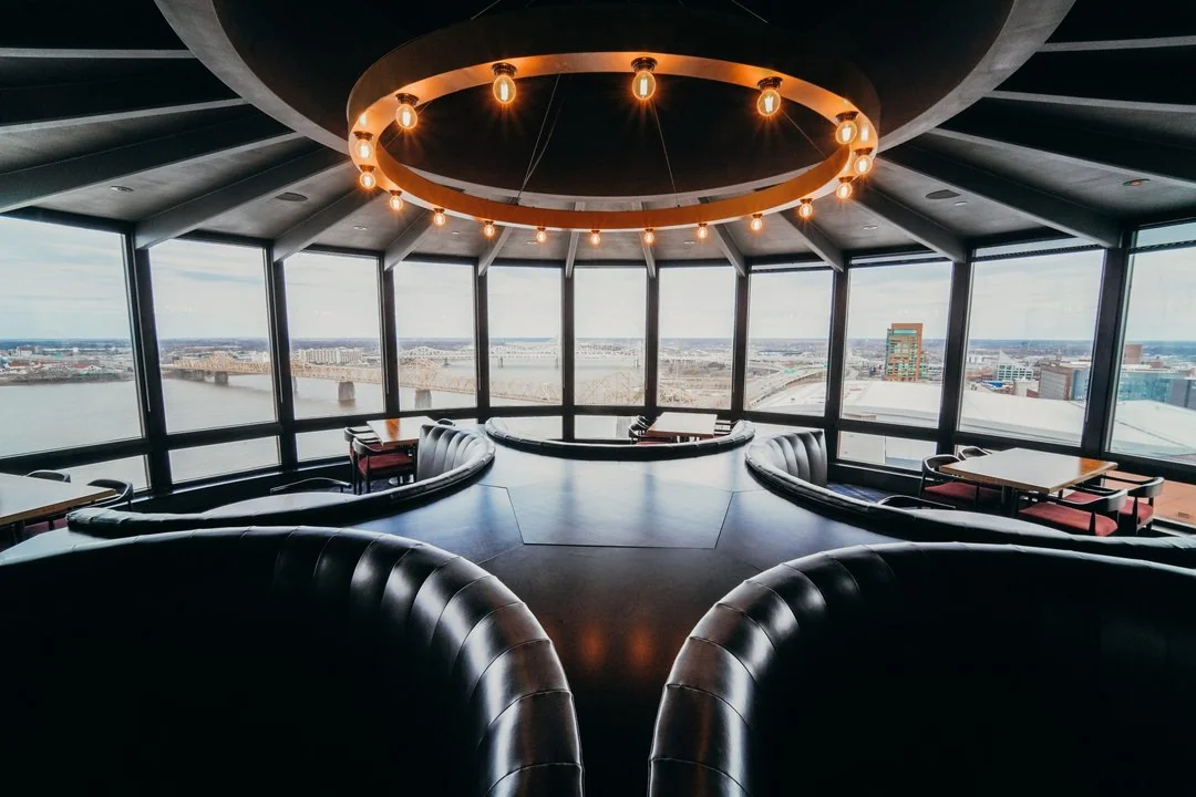 Modern lounge with circular booths and large windows overlooking a city skyline and river. Prominent round chandelier in ceiling. The filename suggests the image is scaled for web use and under 100KB in size.