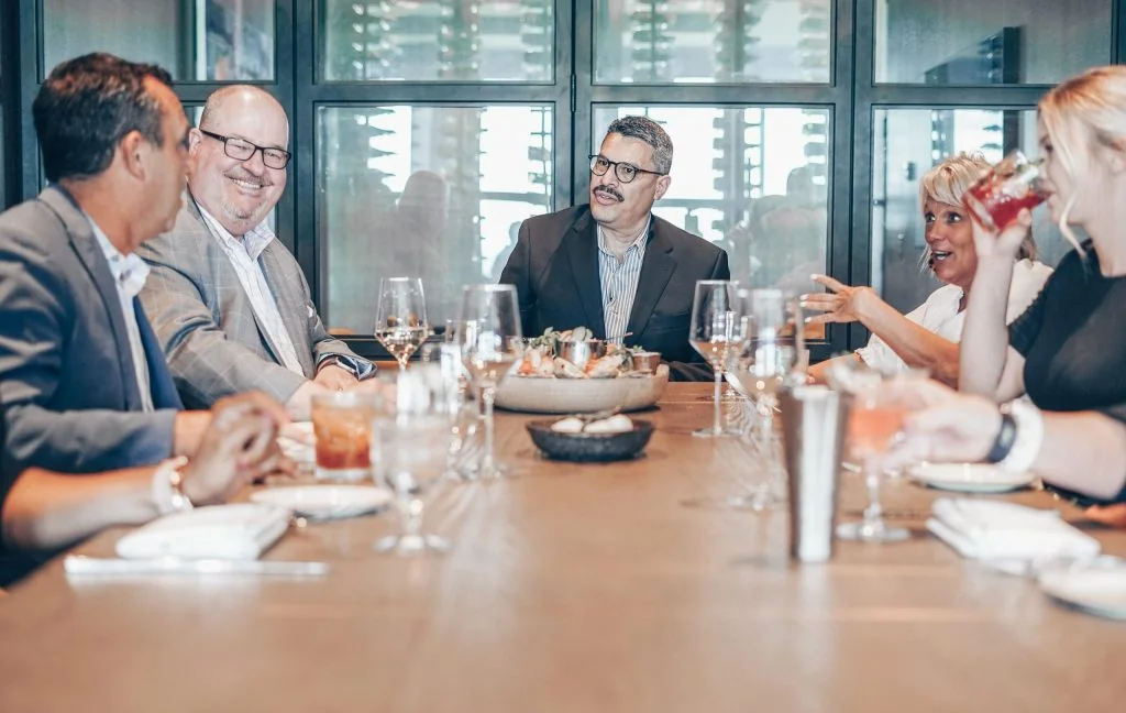 A group of professionally dressed adults sits around a long table, engaged in conversation and drinks, suggesting a business lunch or meeting in a modern restaurant setting.