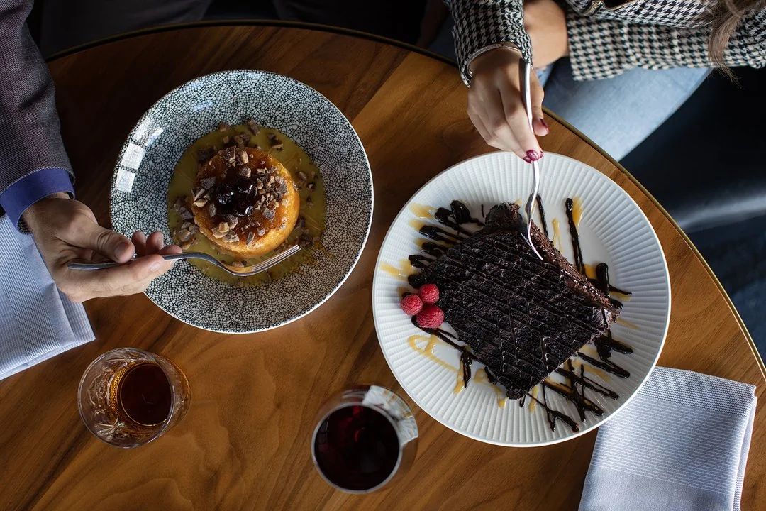 Two people enjoy plated desserts—one has a chocolate cake with raspberries, drizzled with sauce; the other a round cake with nuts and sauce. Drinks are set beside each plate on a restaurant table.