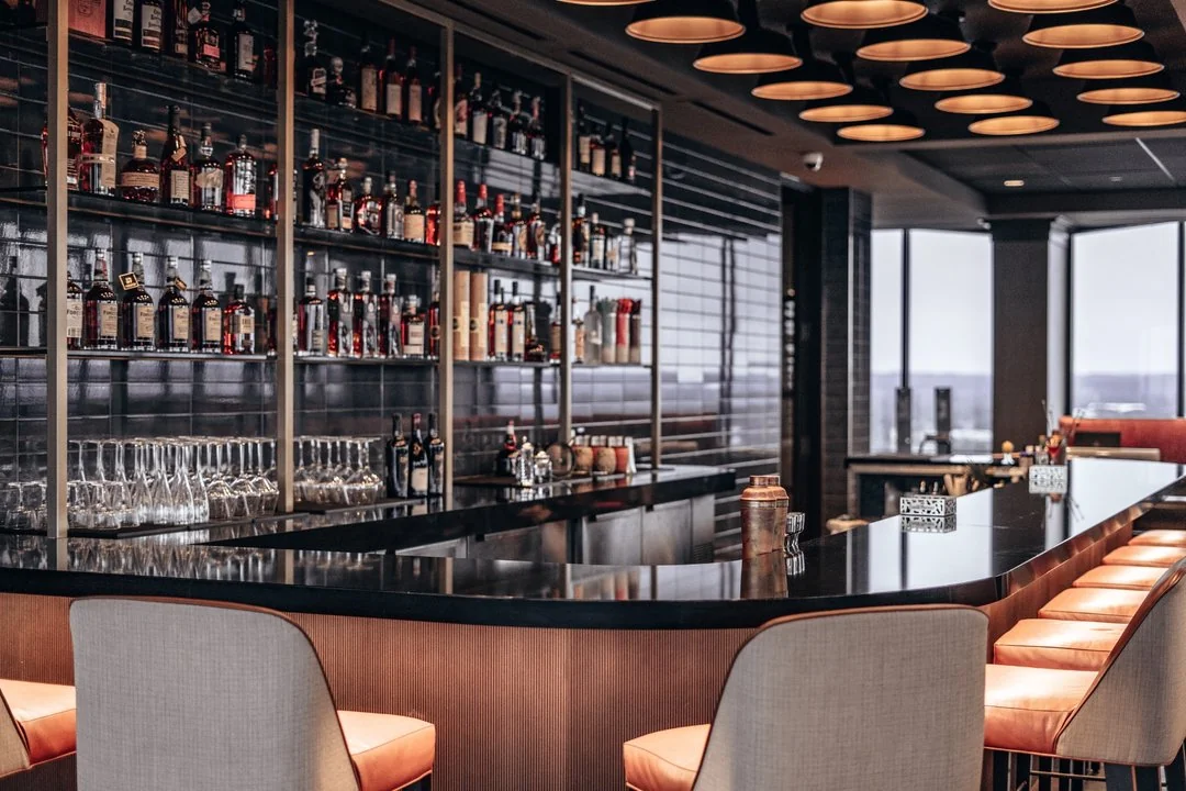 Sleek modern bar with shelves of liquor bottles and glassware, cushioned bar stools, and a glossy black counter. The space has large windows and an upscale, inviting ambiance.